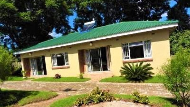 A two-story house with a green roof, a front porch, and a small pond. - 2 Bedroomed Apartment With En-suite and Kitchenette - 2066 (Harare)