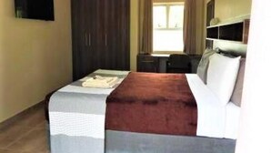 A hotel room with a bed, a desk, a chair, a television, and a window with curtains. - 2 Bedroomed Apartment With En-suite and Kitchenette - 2066 (Harare)