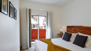 A hotel room with a bed, a nightstand, and a view of a red building through the window. - Ribeira das Casas Apt 2C by Homie (Funchal)