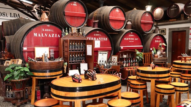 A wine cellar with wooden barrels, shelves of wine bottles, and round wooden tables and stools. - Ribeira das Casas Apt 2C by Homie (Funchal)