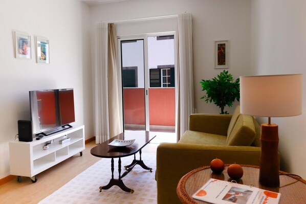 Un salon comprenant un canapé, un téléviseur posé sur un meuble, une table avec une lampe, et une porte-fenêtre coulissante donnant sur l’extérieur.