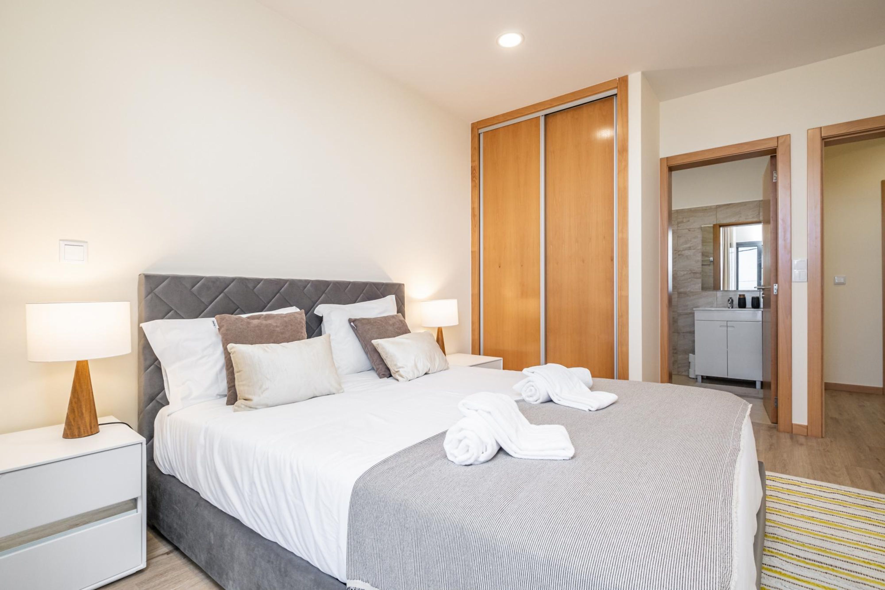 Cama bem arrumada com lençóis brancos, cabeceira cinza e manta cinza. Duas mesas de cabeceira com abajures e uma pequena gaveta. Um guarda-roupa de madeira é visível ao fundo.