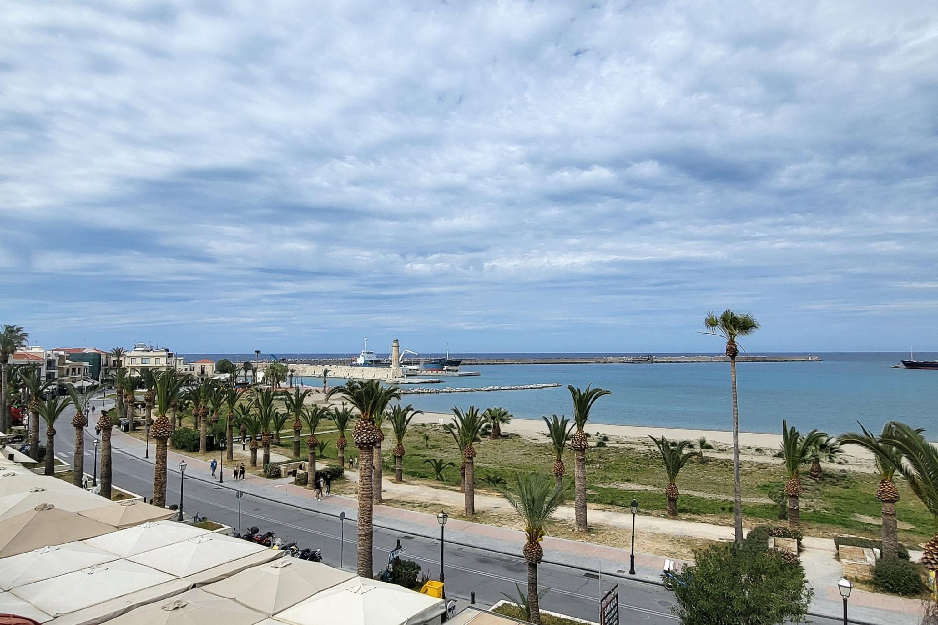 Vista costera con un paseo marítimo bordeado de palmeras, una playa y un faro a lo lejos.