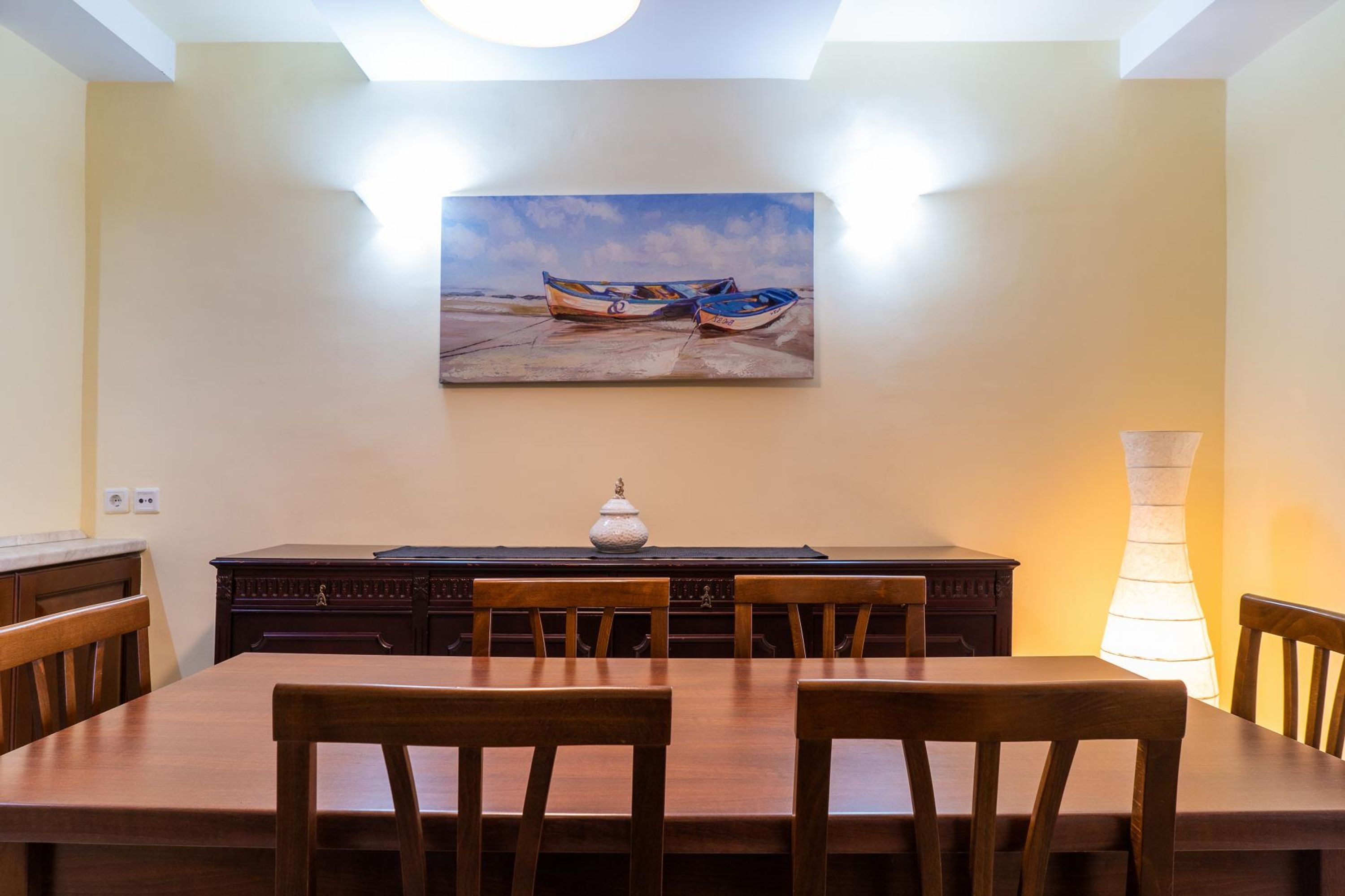 Un coin repas avec une table et des chaises en bois, un tableau représentant des bateaux accroché au mur et un vase posé sur la table.