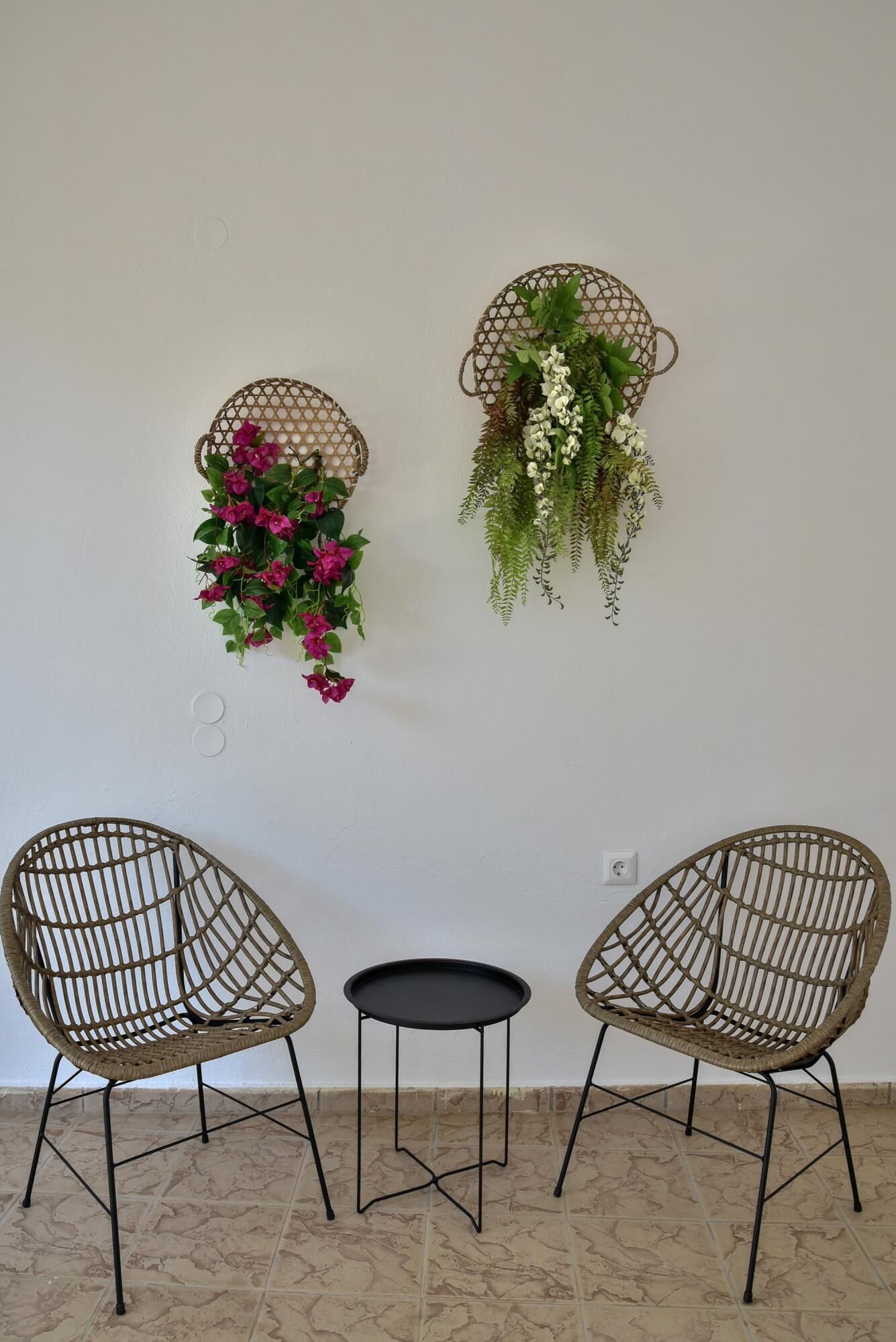 Two wicker chairs with a small black table between them, a white wall, and two wall-mounted planters with flowers.