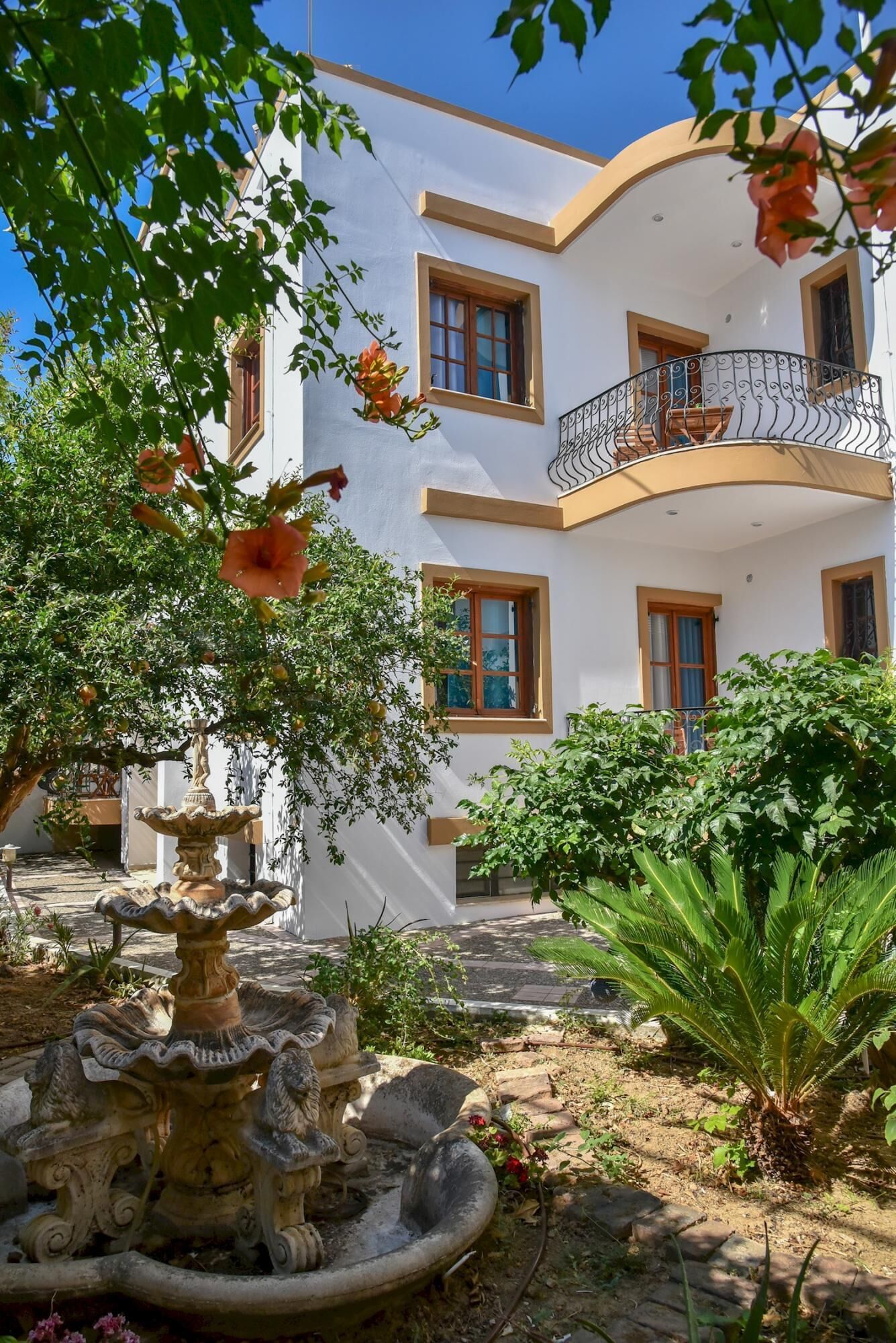A white building with multiple balconies, a fountain, and lush greenery.
