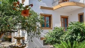 A white building with multiple balconies, a fountain, and lush greenery. - Apartments for 3 2nd Floor (Malevizi)