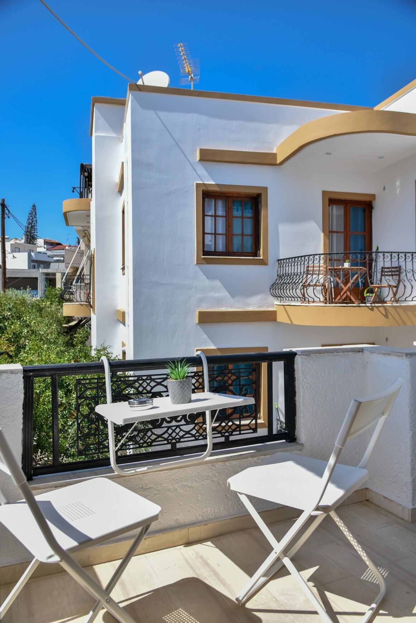 A balcony with white chairs and a table, a potted plant, and a view of a white building with balconies.