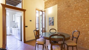 A dining area with a round glass table and wooden chairs, a brick wall, and a view into the kitchen. - Verdi 11 - Two Bedroom Apartment, Sleeps 4 (Firenze)