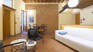 A cozy room with a brick wall, wooden ceiling beams, a white sofa, a small table with chairs, and a door leading to another room. - Verdi 11 - Two Bedroom Apartment, Sleeps 4 (Firenze)