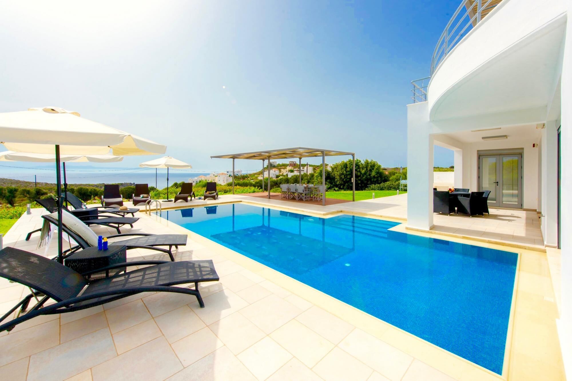 A modern outdoor pool area with lounge chairs and umbrellas, a covered seating area, and a view of the sea.
