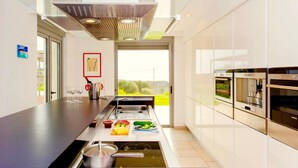 A modern kitchen with a central island, built-in ovens, and a large window offering a view of greenery. - Luxury Villa Stella With Private Swimming Pool (Chania)