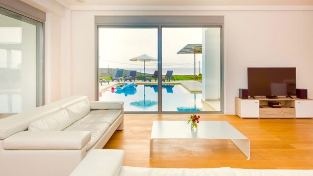 A modern living room with a white sofa, a coffee table, and a TV on a wooden stand. A large sliding glass door leads to an outdoor pool area with lounge chairs and umbrellas. - Luxury Villa Stella With Private Swimming Pool (Chania)