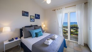 A bedroom with a bed, two bedside tables, a ceiling fan, and a balcony with a view of the sea. - Sirina Waterfront House (Paralimni)