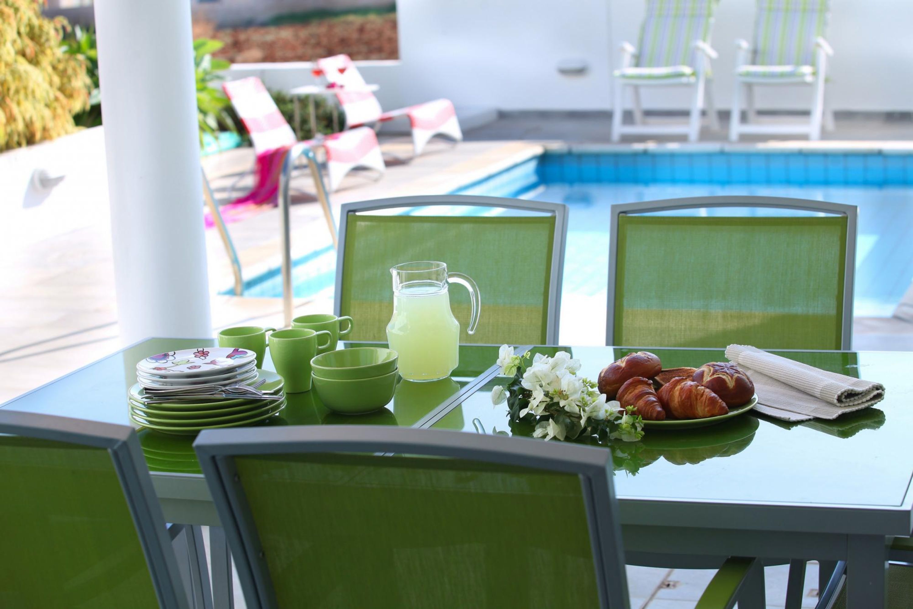 A dining table with green chairs, a pitcher of lemonade, plates, cups, and a plate of croissants.