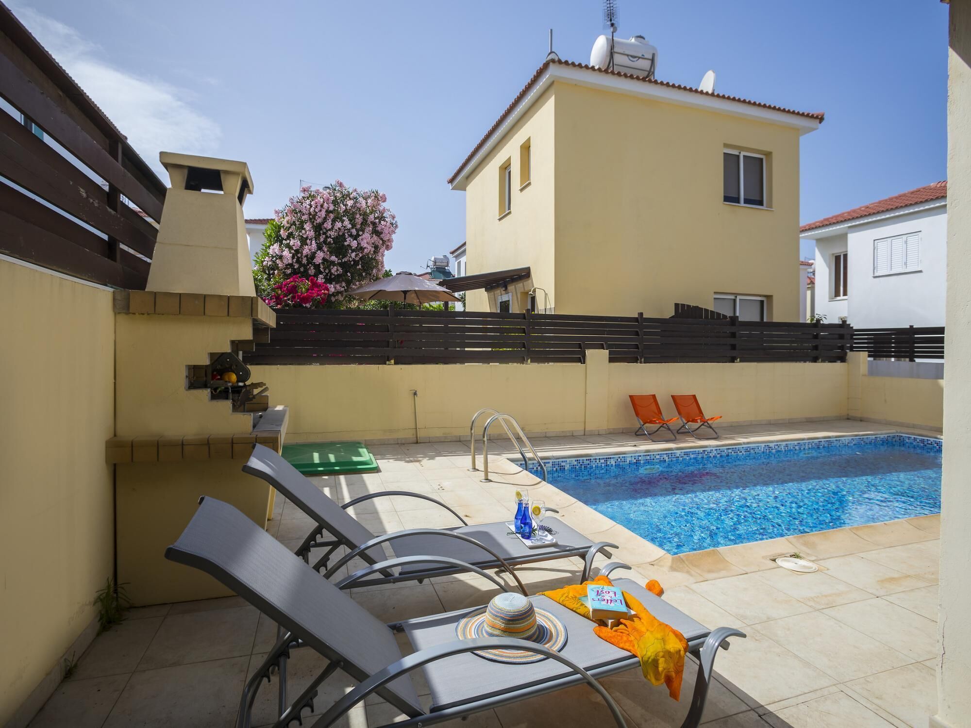 A pool area with lounge chairs, a small table with a bottle and a hat, and a flower bush.