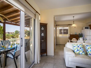 Amplia sala de estar con puerta corrediza de vidrio que da a un patio exterior, un sofá blanco con cojines florales y una zona de cocina con refrigerador y microondas.