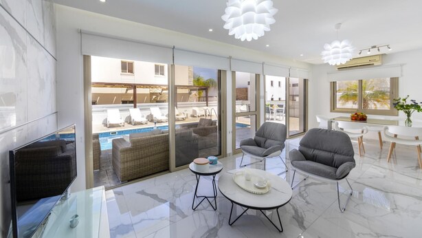 Een moderne woonkamer met een glazen salontafel, grijze fauteuils, een bank en uitzicht op een zwembad.