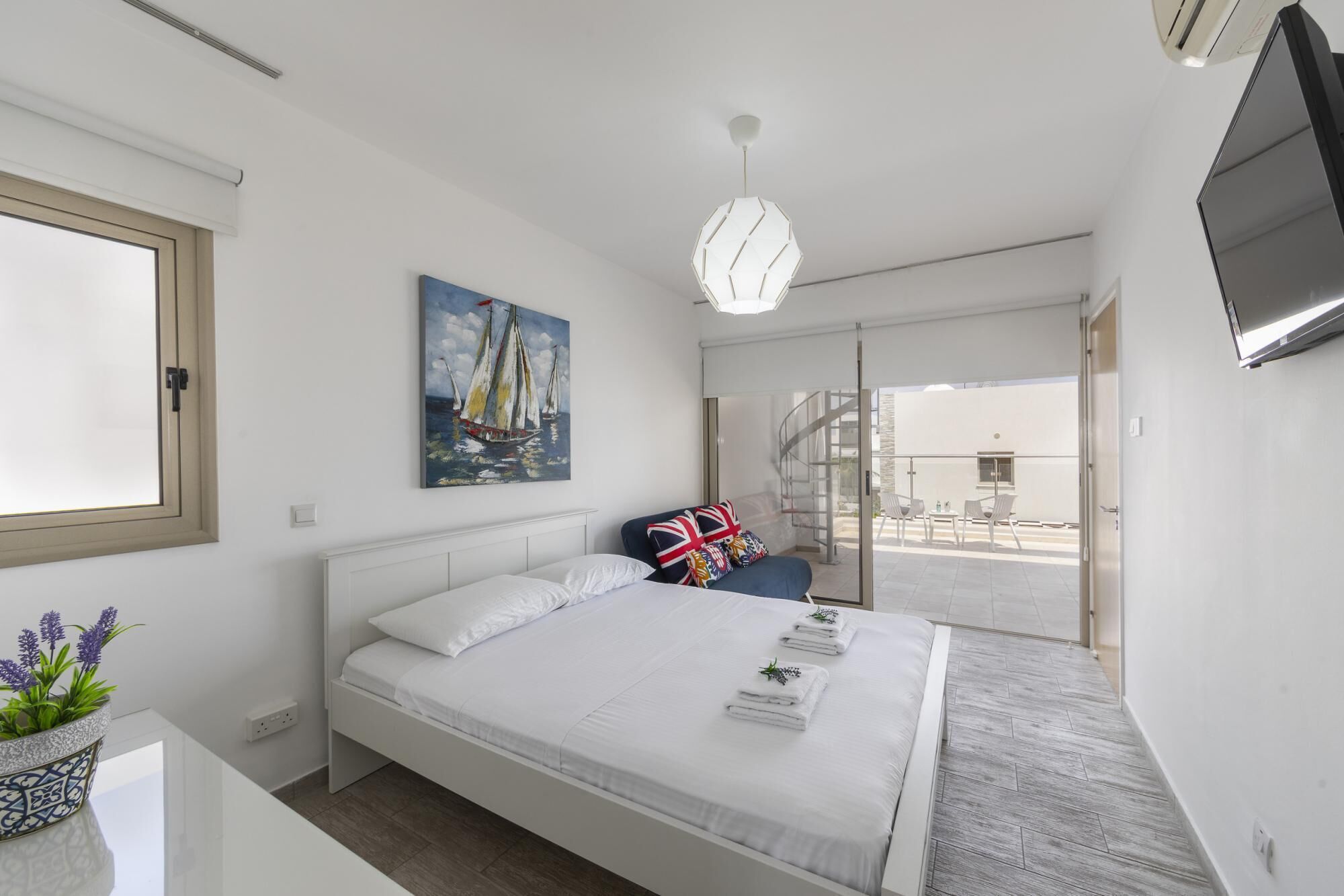 A modern bedroom with a large bed, a painting of sailboats, a TV, and a balcony with a spiral staircase.