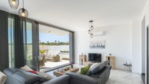 A modern living room with a grey sectional sofa, a flat-screen TV, and a large sliding glass door leading to an outdoor patio. - Protaras Royal Pearl Villa 7 (Paralimni)