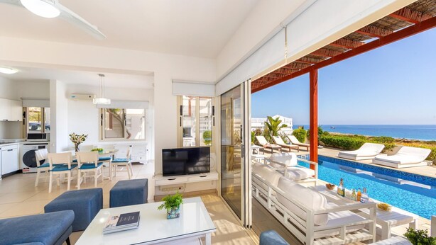 A modern living room with a TV, seating, and a view of a pool and ocean. - Protaras Artisan Seafront Villa 6 (Paralimni)