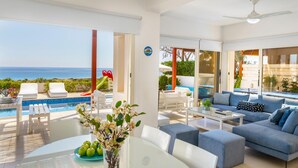 A modern living room with a view of the sea, a pool, and outdoor seating. - Protaras Artisan Seafront Villa 6 (Paralimni)