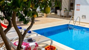 Zona junto a la piscina con pileta, sillas de descanso, una mesa con un sombrero y un cuenco de frutas, y un árbol frutal.