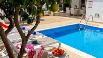 A poolside area with a swimming pool, lounge chairs, a table with a hat and a bowl of fruit, and a tree with fruit.
