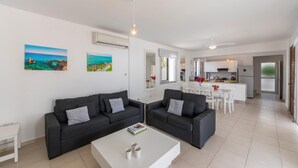 Un moderno salón con dos sofás negros, una mesa de centro blanca y una zona de comedor con una mesa y sillas blancas. Hay dos cuadros en la pared, una cocina al fondo y un ventanal grande.