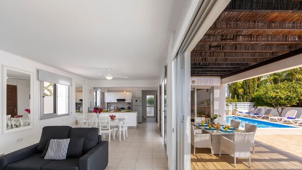 Una sala de estar moderna con un sofá negro, un comedor con mesa y sillas, una cocina al fondo y una zona de piscina con sillones de descanso.