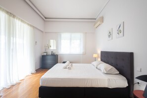 A bedroom with a large bed, a dresser, a window with sheer curtains, and a wall-mounted air conditioner.