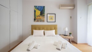 Un lit bien fait, avec du linge de lit et des serviettes blanches, une table de chevet avec une lampe et une petite plante, un tableau encadré au mur et une grande fenêtre avec des rideaux.