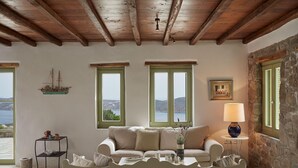 A cozy living room with a sofa, a small table, and a lamp. There are windows with a view of the sea and mountains. - Spacious 6 Bdrm Stone Villa With Pool (Serifos)