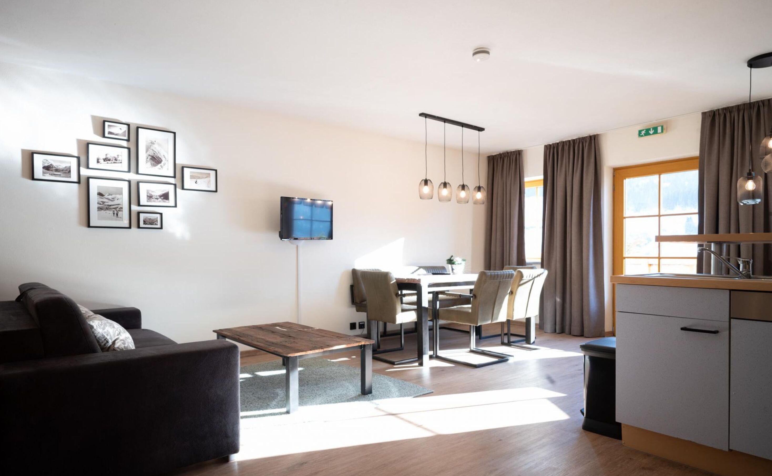 A modern living room with a sofa, a coffee table, and a TV mounted on the wall. There is a dining area with a table and chairs, and a kitchen area with a sink and cabinets.