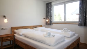 A double bed with white linens and towels, a wooden nightstand, and a window with curtains. - Tauern Relax Lodges - Penthouse (Kaprun)