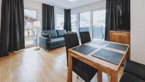 Ein modernes Wohnzimmer mit einer Couch, Sesseln, einem Holztisch und Blick auf eine Berglandschaft durch große Fenster.