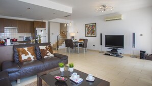 Una sala moderna con un sofá de cuero, una mesa de centro y un televisor de pantalla plana. Se ve un comedor con una mesa y sillas al fondo.