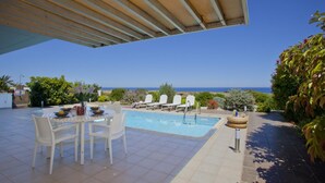 A pool area with a view of the ocean, a covered patio with a table and chairs, and a small garden with various plants. - Villa Melissa Selana (Paralimni)