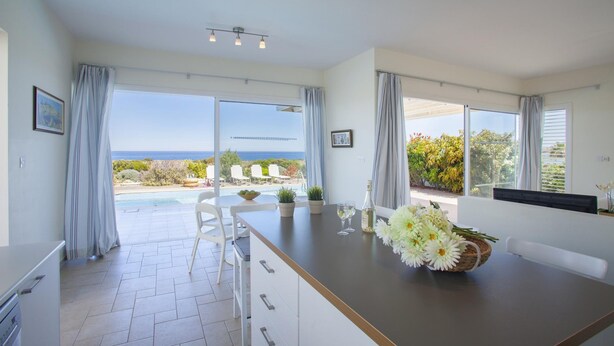 A modern kitchen with a view of the ocean, a dining area with a table and chairs, and a large window with curtains. - Villa Melissa Selana (Paralimni)