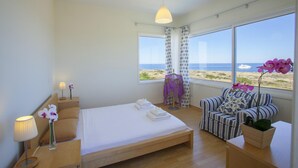 A bedroom with a bed, a chair, a nightstand, a lamp, and a view of the sea. - Villa Melissa Selana (Paralimni)