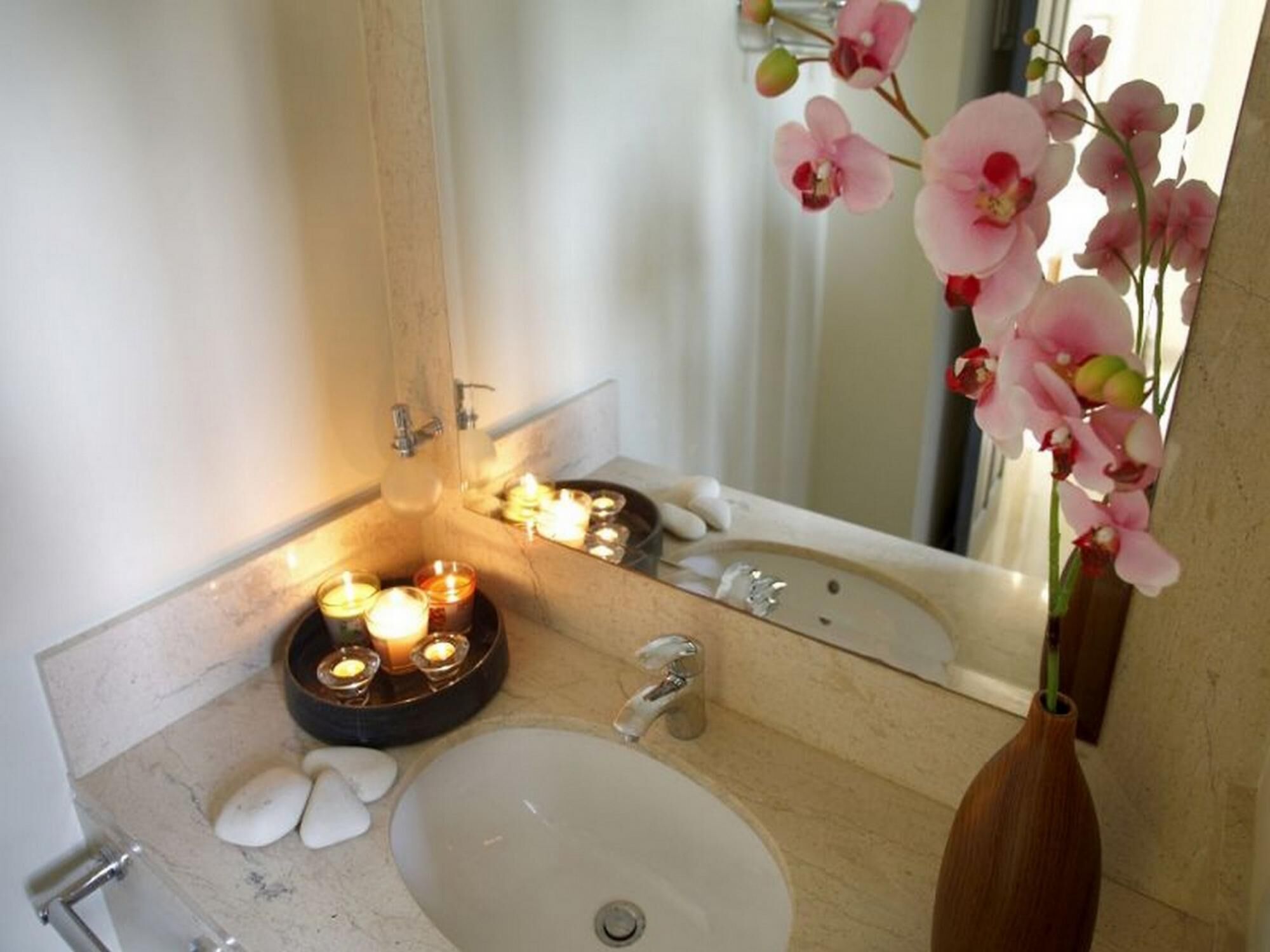 A bathroom with a white sink, a mirror, and a vase with pink orchids. There are candles on the counter and a wooden bowl with stones.