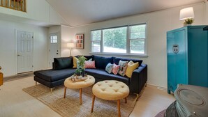 A living room with a dark-colored sectional sofa, two round ottomans, a teal cabinet, and a large window providing natural light. - Maryland Vacation Rental w/ Private Pool & Dock (Lusby)
