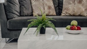 Ein modernes Wohnzimmer mit einem grauen Sofa, einem weißen Couchtisch, einem Farn im Topf und einer Schale mit Obst.
