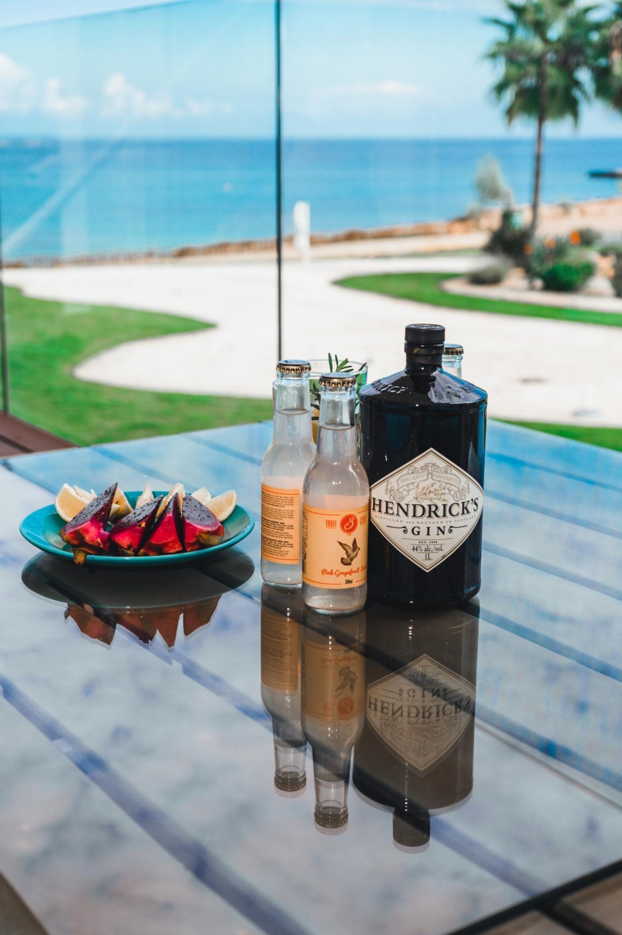 Una mesa con bebidas y fruta junto a un ventanal con vistas a una playa.