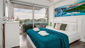 Una habitación de hotel moderna con una cama grande, vista a un paisaje montañoso a través de un ventanal corredero, y una mesita de noche blanca con un teléfono y una botella verde.