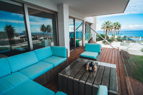 A balcony with a turquoise sofa and a glass coffee table, overlooking a beach with palm trees and the ocean.