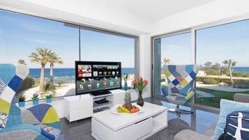 Uma sala de estar moderna com uma televisão grande, uma mesa de centro branca com frutas, e vista para o oceano e palmeiras.