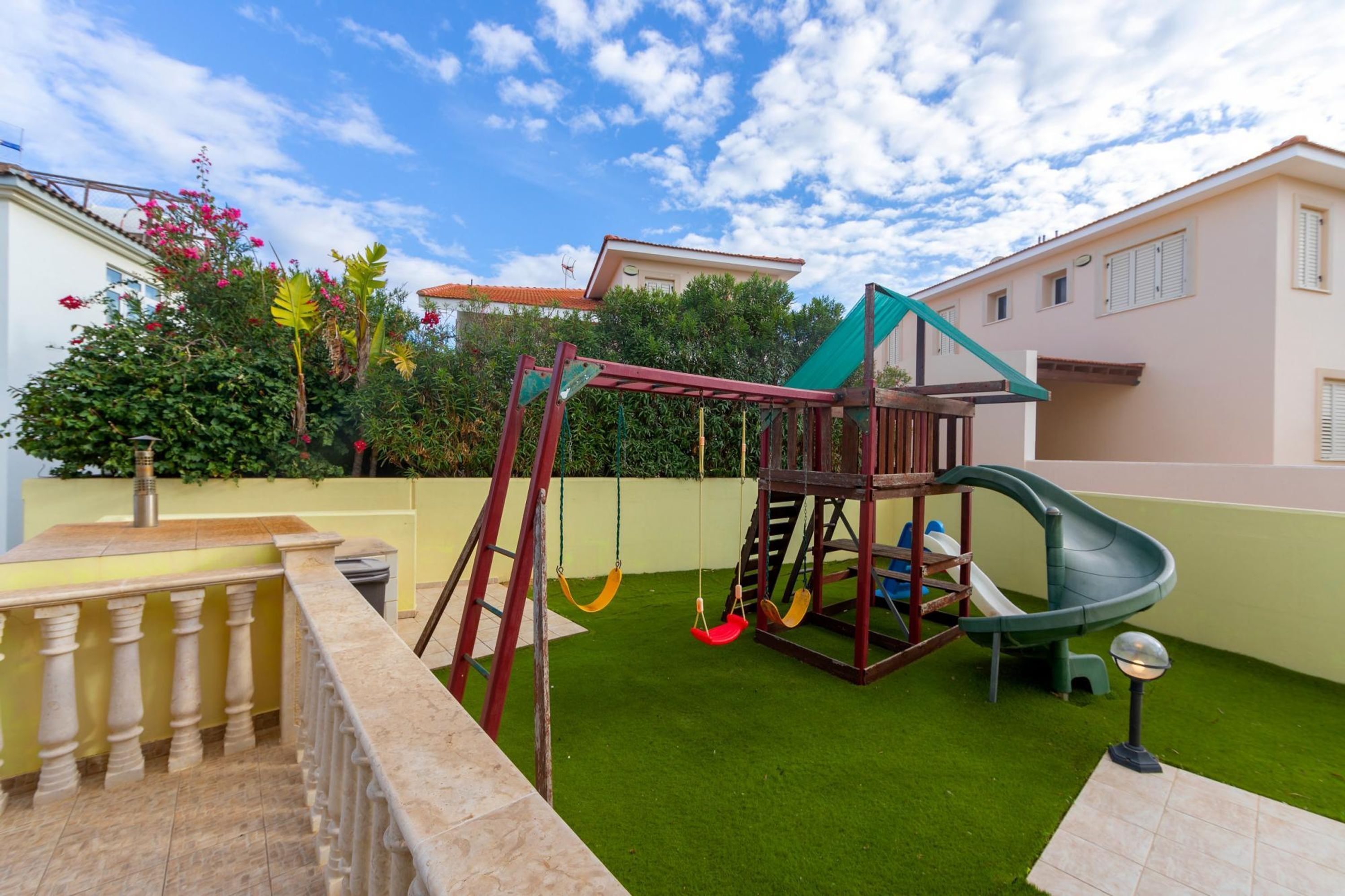 A playground with swings and a slide, surrounded by a green lawn and a beige wall.
