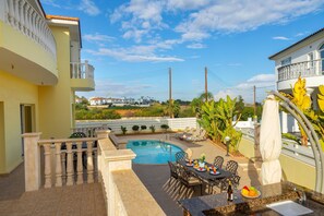 A rooftop pool area with a dining table and chairs, surrounded by a white fence and lush greenery.