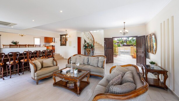A spacious living area with a sofa set, coffee table, and wooden chairs. A staircase leads to a balcony with a view of the outdoors. - Protaras Posidon By The Sea (Paralimni)